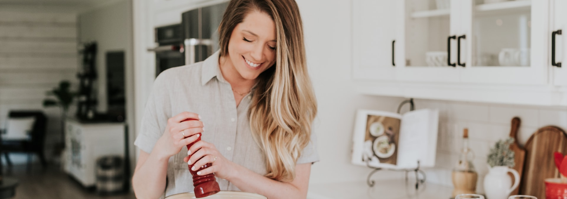 Uma mulher está na cozinha de sua casa, um ambiente integrado, com um moedor de pimenta nas mãos e sorri.